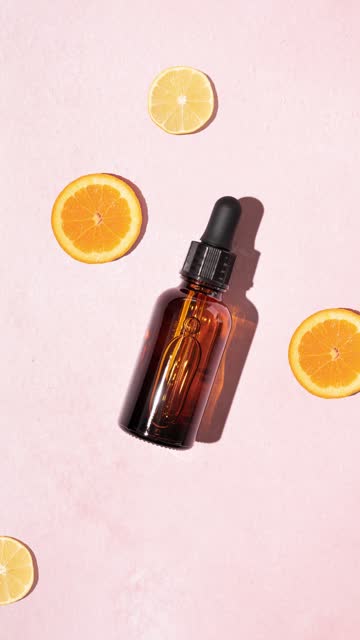 Vivid fresh vitamin C serum with brown glass cosmetic dropper bottle and slices of lemon and orange on a pastel pink background. Natural bright skincare concept.