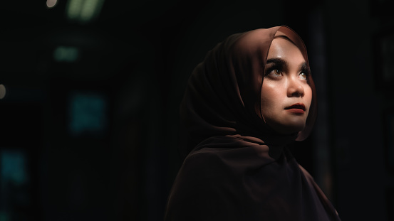 A Muslim woman in a Muslim veil hijab standing in a dark room and looking away.