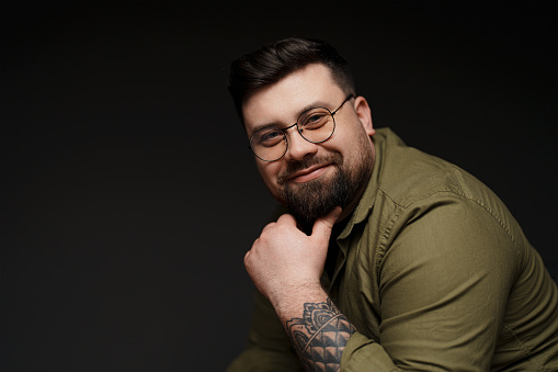 Thoughtful young plump tattooed male model in eyeglasses and green shirt looking at camera and touching beard against black background in studio