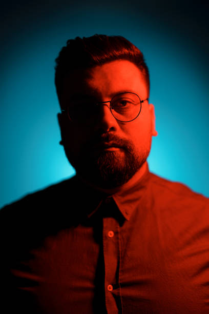 Portrait of unemotional young bearded male model wearing shirt and glasses looking at camera in red neon illumination background with shadow in studio