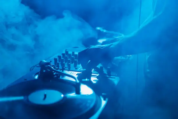 Hands of a disc jockey playing music on party in night club in bright blue lights and thick smoke