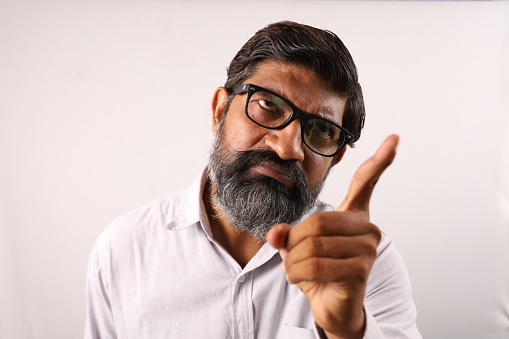 White background conceptual studio shoot of a furious Indian bearded man pointing his Index finger towards the camera.