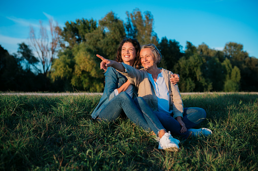 Portrait of beautiful young woman sitting her mother pointing at distance in nature. Loving mother and daughter sitting on grass lawn outdoors.