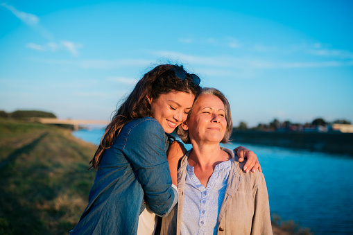 Affectionate woman with her mother standing by river. Loving daughter with her mother bonding together outside in nature on mother's day.