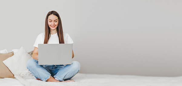 freelancer girl sits on a bed using laptop, looks at the monitor and works web banner. Girl student studying remotely at home. Online learning English. copy space