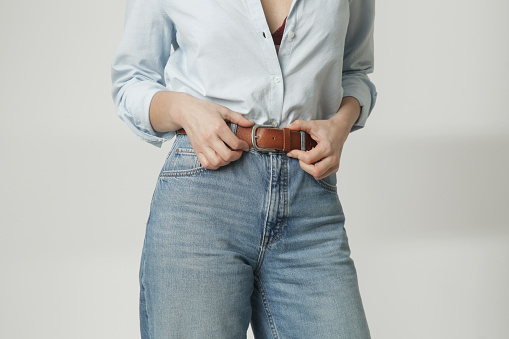 Fashion portrait of young woman in cotton button down and blue jeans adjusting her brown leather belt