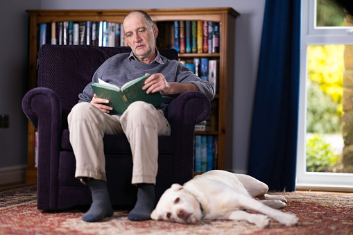 Caucasian man in his 60s reading a book in his study with his faithful dog by his side.