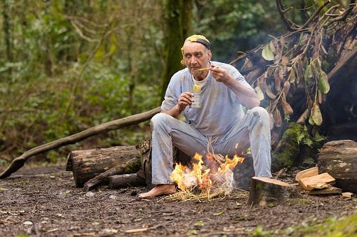 Homeless man in his 60s sitting by his pathetic fire and bivouac eating a can of cold soup.