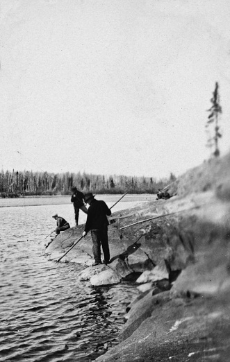 Wekusko Lake, Manitoba, Canada - 1924. Three men fishing with rods and spear on the edge of Wekusko Lake in Manitoba, Canada. Vintage photograph ca. 1924.