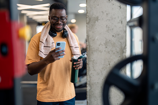 Handsome African American sportsman using mobile phone while taking a break from exercising, standing in gym and smiling. Young fit male fitness instructor texting on mobile phone while resting in gym.