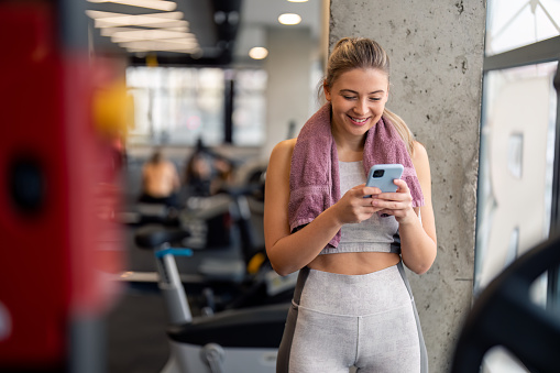 Fit young woman using mobile phone in gym to watch virtual workout tutorial video to improve her fitness workout exercise. Motivated sportswoman watching online fitness tutorial on smartphone in gym.