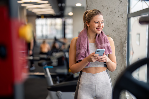 Happy athletic woman using phone while taking a break from exercising, standing in gym looking out the window and smiling. Beautiful young female gym member texting on mobile phone while resting in gym.