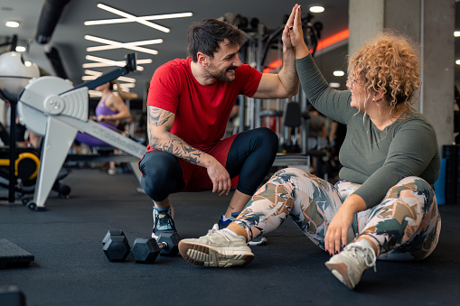 Happy male coach giving young woman high-five after successful sports training session in gym. Two friends at gym cheerful man and woman giving each other high-five after hard workout celebrating success.