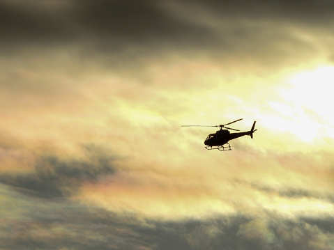 An Aerospatiale AS350 helicopter flies across the flightpath of Sydney Kingsford-Smith Airport at sunset.  This image was taken from Shep's Mound, a public viewing area off Ross Smith Avenue, Mascot on a cloudy and overcast day at sunset on 1 April 2023.
