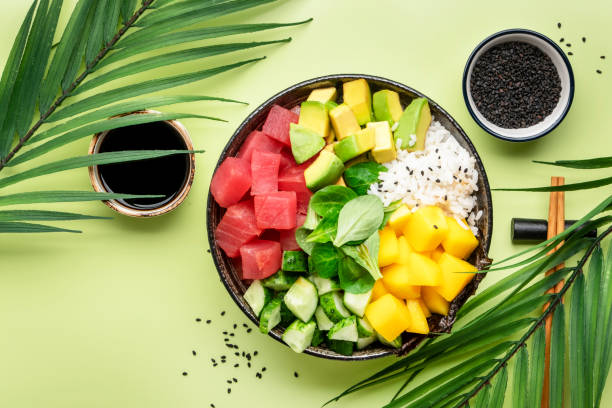 Poke bowl with raw tuna, avocado, mango, cucumber, lettuce and white rice. Soy sauce, lime and sesame dressing. Green table background, top view stock photo