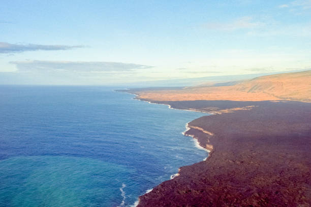 A vintage 1980s film photograph aerial view of the Hawaiian volcanic coastline where the lava meets the sea from a helicopter on a clear blue sky day.
