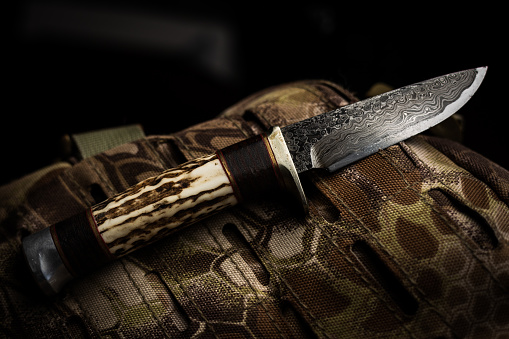 A hand made custom damascus blade with deer antler handle hunting knife resting on a camouflage tactical backpack.