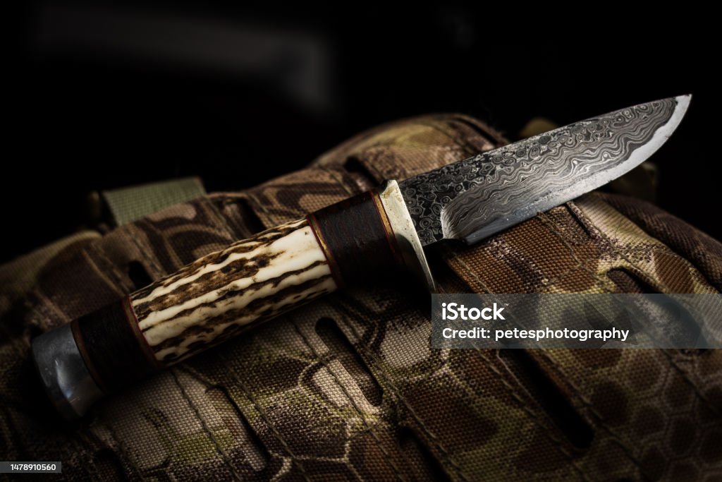 A custom Damascus and deer antler hand made knife A hand made custom damascus blade with deer antler handle hunting knife resting on a camouflage tactical backpack. Advanced Tactical Fighter Stock Photo