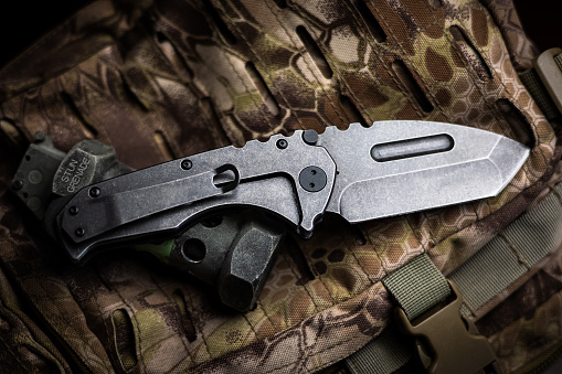 A tactical tanto style full steel folding pocket knife displayed on a flashbang grenade and camouflage backpack.