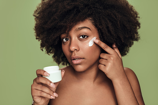 Young black female with bare shoulders and Afro hairstyle looking at camera and smearing moisturizing cream on cheek against green background