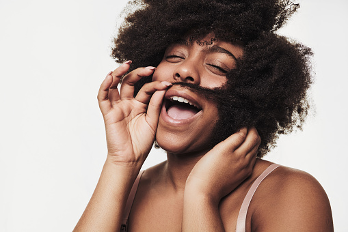 Cheerful young African American female model laughing with closed eyes while making mustache with curly dark hair against white background