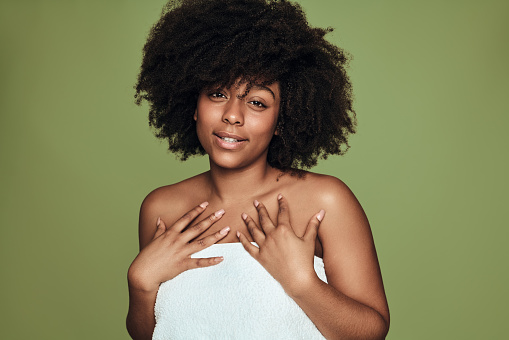 Young African American female wrapped in white towel with curly hair touching chest and looking at camera during skin care routine against green background