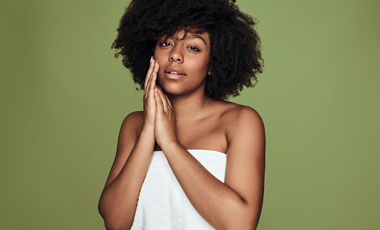 Young African American female with Afro hairstyle wearing towel after shower gently touching face and looking at camera on green background