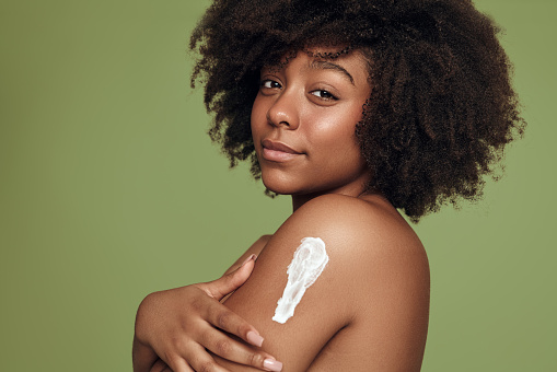 Side view of young black female with Afro hairstyle applying body lotion on arm and looking at camera during spa session against green background