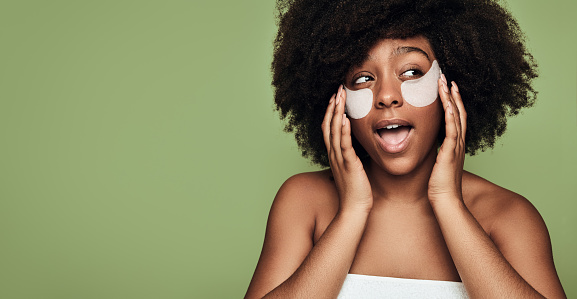 Positive confident young black female millennial with Afro hair applying hydrating collagen patches on face and looking away with opened mouth against green background