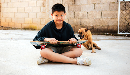 Smile Asian boy sitting on a skateboard. Outdoor portrait of lovely Asian kid boy model posing with brown dog