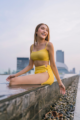 summer travel relax vacation portrait asian woman swimwear sit on pool side order cheerful happiness smiling positive pleasure expression at building roof top infinity pool with building background