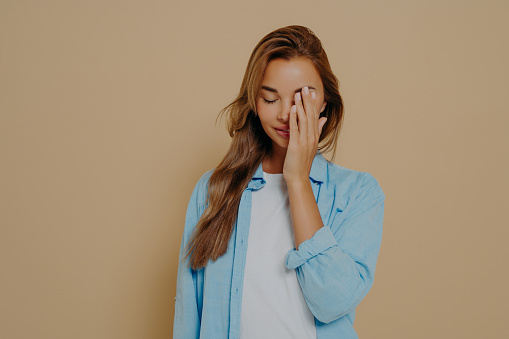 Portrait of tired and annoyed young female hiding eyes as being embarrassed making facepalm gesture tilting head down standing upset and irritated over beige background in blue oversized shirt