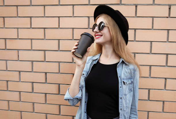 jeune femme élégante avec la tasse de café regardant loin utilisant un chapeau rond noir, veste de jeans sur le fond de mur de brique - back against the wall photos et images de collection