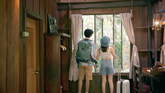 Rear view of happy young Asian couple, enjoying vacation together by traveling to countryside and standing, looking through window with view at amazing mountains, forest in wooden house, summer travel concept. Cabin getaway.
