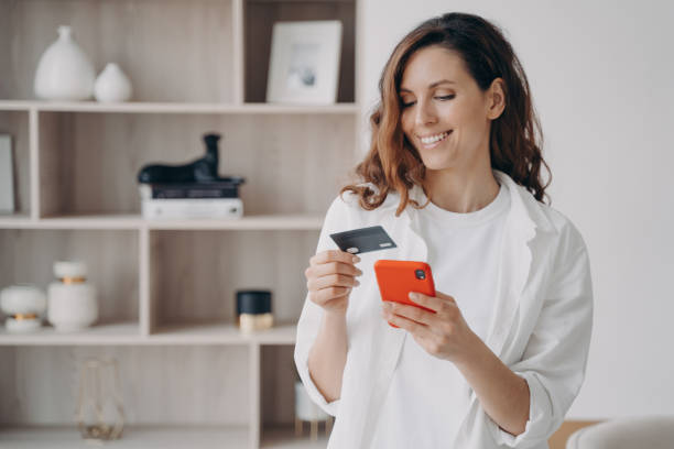 Easy payment with smartphone and card. Happy caucasian girl is going to buy through internet. Easy payment with smartphone and card. Happy caucasian girl is going to buy through internet at home. Girl is booking or purchasing online. E-commerce and financial transaction concept. paying stock pictures, royalty-free photos & images