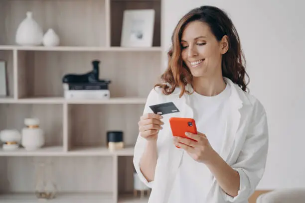 Easy payment with smartphone and card. Happy caucasian girl is going to buy through internet at home. Girl is booking or purchasing online. E-commerce and financial transaction concept.