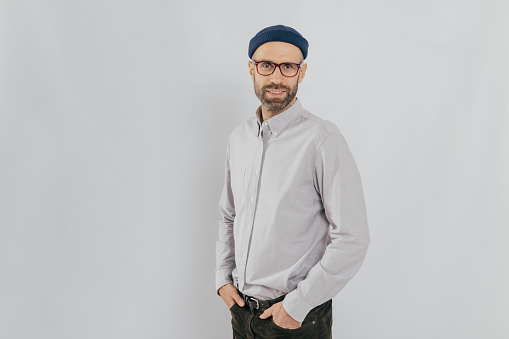 Sideways shot of handsome pleased man wears stylish hat, keeps hands in pockets, stands against white background with copy space for your advertisement or promotion. Hipster guy models indoor