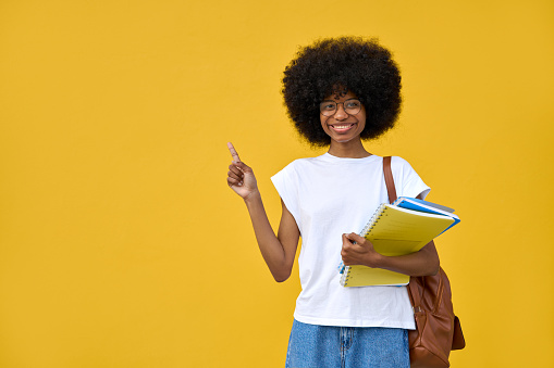 Young happy african american smart student girl with curly afro hairstyle stand with backpack and hold exercise books, showing on copy space for advertising and looking at camera on yellow background.