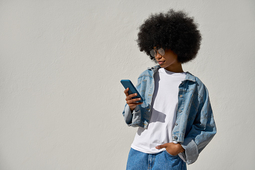 A hipster girl with afro haircut enjoy online communication and online services, wear trendy jeans jacket, scroll social media apps on cell phone device isolated with copy space on white background