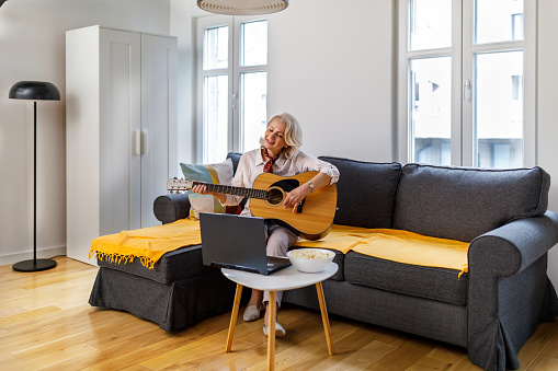 An older woman is practicing playing an acoustic guitar while sitting on a sofa in a comfortable apartment and watching tutorials on a laptop. Musical hobbies concept.