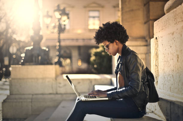 istock Young black female with a black jacket concentrated on work with her laptop while sitting outside 1479019211