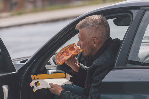 Adult man is eating pizza outdoors inside of parked car