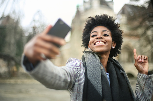 A young black female with a grey coat taking a selfie with her phone outside