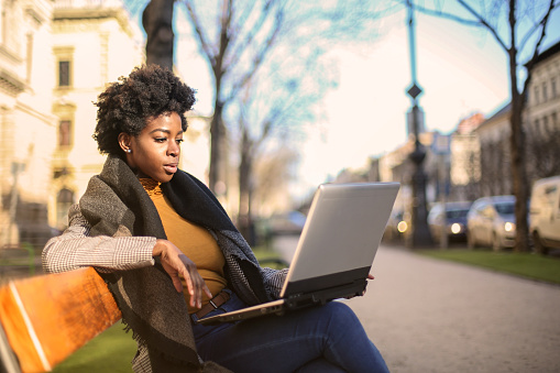 A young black female sitting on a bench outside and working on her laptop