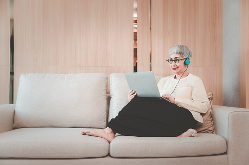 Senior mature grey hair woman in eyeglasses and headphone sitting on couch using computer laptop having online meeting, Aging society concept