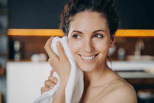 Evening beauty routine and hygiene. Caucasian woman is wiping face with towel after washing. Young happy woman takes shower at home. Face and body care, dermatology and spa procedures.