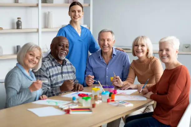 Cheerful attractive young woman in workwear nurse posing with multiracial group of happy senior people while having art-therapy class at assisted living, elderly men and women enjoying painting