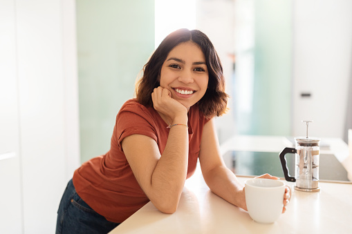 Beautiful Young Middle Eastern Female Drinking Morning Coffee In Kitchen At Home, Happy Millennial Arab Woman Standing At Counter And Smiling At Camera, Enjoying Fresh Caffeine Drink, Free Space