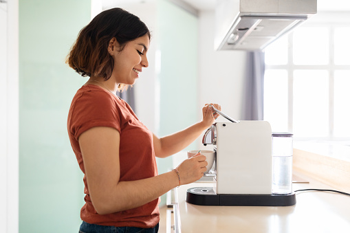 Young smiling arab woman preparing fresh coffee with modern machine in kitchen, side view shot of beautiful middle eastern female cooking caffeine drink in the morning at home, copy space