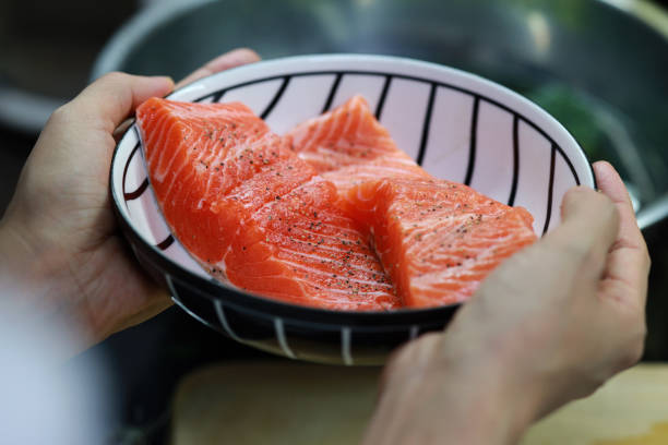 Cropped shot of woman marinated raw salmon fillet with salt and pepper in kitchen